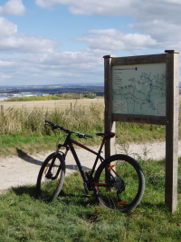 Bicycle leaning against Ridgeway long distance path information board, with the track running across the image in front.  On the far side, across a corn field, is the ERU Test Site, on the south side of STFC Rutherford Appleton Laboratory.  Above are blue skies and white clouds.
