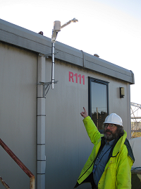 Portacabin labelled R111, with Temperature Probe, Pyranometer and Humiidity Probe  mounted on a pole projected a short distance above the roof.  Peter, in hard hat and high visibility jacket, points at the guages.